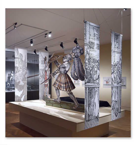 Ausstellungsschaubild im Reiss-Engelhorn-Museum – zwei kämpfende Ritter umrahmt von 4 von der Decke hängenden Spannbändern, auf denen das Publikum dazu abgebildet ist.